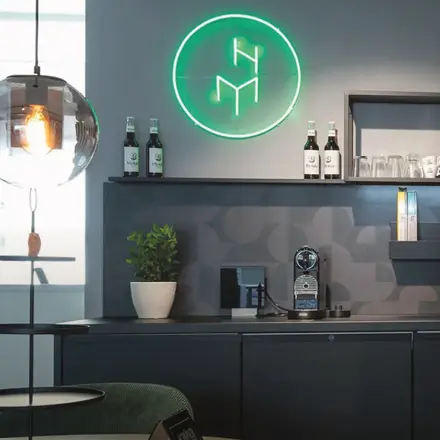 Dunkle Kaffeebar mit Unterschränken, weissem Blumentopf, Kaffeemaschine und Wandregalen, darüber rundes leuchtendes Logo mit den Buchstaben H und M.